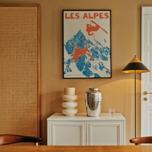 May include: A framed vintage poster with the text "LES ALPES" depicts skiers on a snowy mountain. A white cabinet holds a stacked vase, a silver vase, and a glass. A black and gold floor lamp stands nearby.