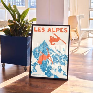 May include: Framed vintage poster with the text "LES ALPES" in orange, depicting skiers against a blue and white mountain backdrop. The poster is in a black frame, leaning against a blue planter with a plant.