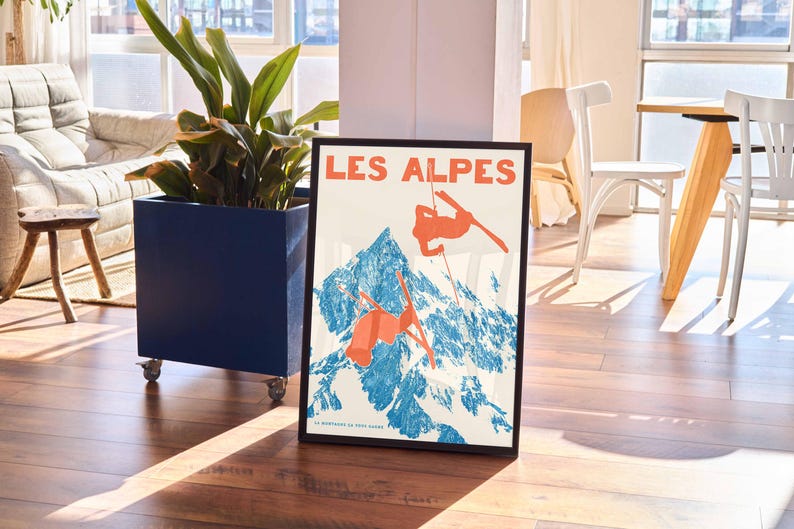 May include: Framed vintage poster with the text "LES ALPES" in orange, depicting skiers against a blue and white mountain backdrop. The poster is in a black frame, leaning against a blue planter with a plant.
