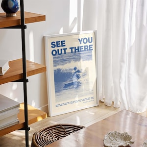 K&ouml;nnte beinhalten: Ein wei&szlig;er gerahmter Poster mit blauem Text, der "See You Out There" lautet, und einer blauen und wei&szlig;en Illustration einer Person beim Surfen. Der Poster h&auml;ngt an einer wei&szlig;en Wand.