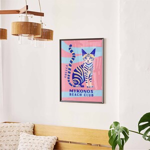 May include: A colorful poster with a blue and yellow striped cat sitting on a pink background. The poster says "Greece" and "Mykonos Beach Club".