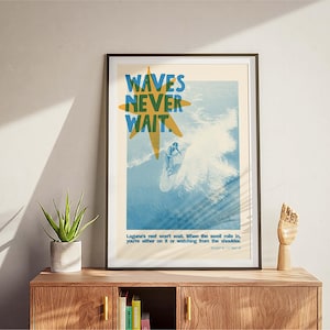 May include: Framed art print with a surfing theme. The poster features the text "WAVES NEVER WAIT" in blue and yellow, with a surfer riding a wave. The print includes additional text at the bottom. The artwork is displayed on a wooden cabinet.