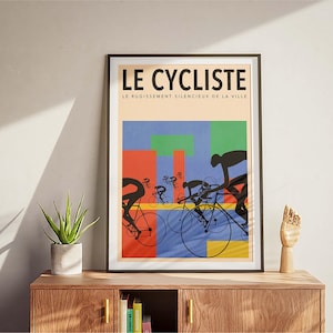 May include: Framed art print titled "LE CYCLISTE" featuring cyclists in silhouette against a colorful geometric background. The artwork includes the text "LE RUGISSEMENT SILENCIEUX DE LA VILLE". The print is displayed on a wooden cabinet.