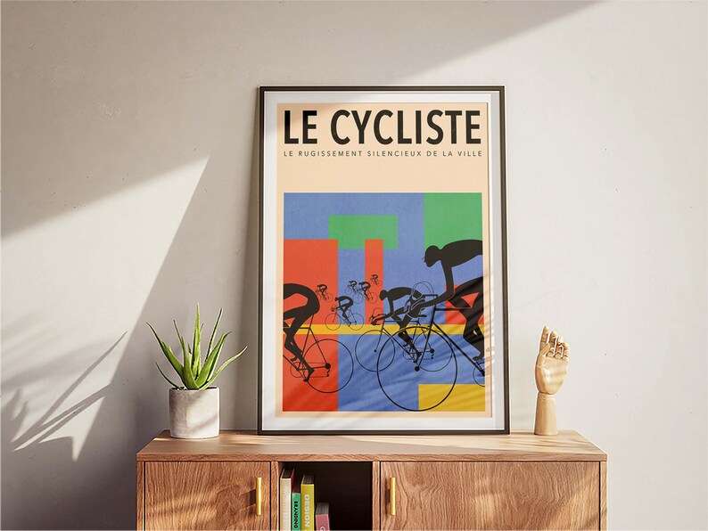 May include: Framed art print titled "LE CYCLISTE" featuring cyclists in silhouette against a colorful geometric background. The artwork includes the text "LE RUGISSEMENT SILENCIEUX DE LA VILLE". The print is displayed on a wooden cabinet.