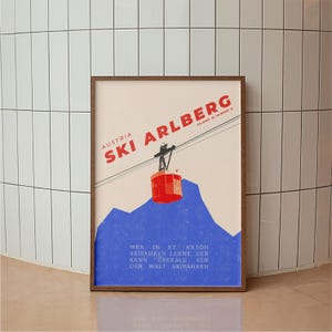 May include: A framed vintage travel poster for Ski Arlberg, Austria. The poster features a red cable car, blue mountains, and the text "SKI ARLBERG" in red. The poster also includes German text at the bottom.