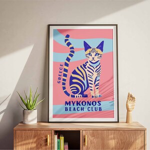May include: A pink and blue poster with a stylized illustration of a cat. The poster has the text "GREECE" and "MYKONOS BEACH CLUB".