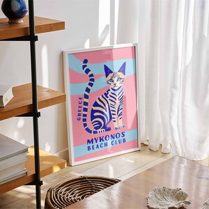 May include: A white framed poster with a pink background and a blue and yellow striped cat sitting on a blue and white striped wave. The text on the poster reads "Greece" and "Mykonos Beach Club".