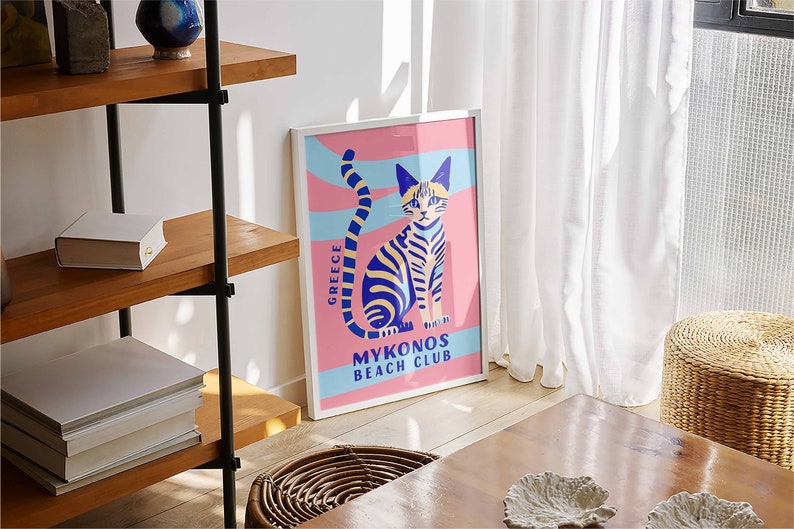 May include: A white framed poster with a pink background and a blue and yellow striped cat sitting on a blue and white striped wave. The text on the poster reads "Greece" and "Mykonos Beach Club".