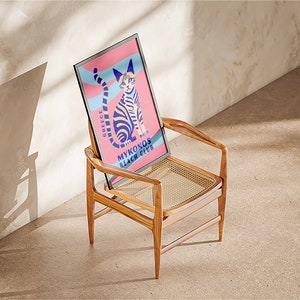 May include: A framed poster with a colorful illustration of a cat on a pink and blue background. The poster says "Greece" and "Mykonos Beach Club". The poster is sitting on a wooden chair with a woven seat.