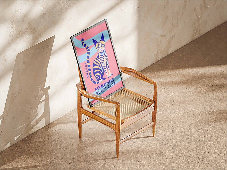 May include: A framed poster with a colorful illustration of a cat on a pink and blue background. The poster says "Greece" and "Mykonos Beach Club". The poster is sitting on a wooden chair with a woven seat.
