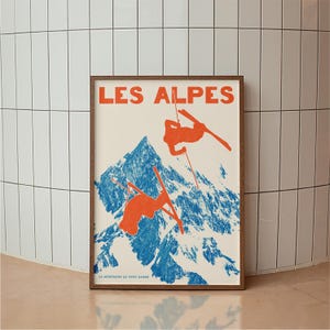 May include: A framed vintage poster with the words "LES ALPES" in orange. The poster depicts a blue mountain and two orange skiers. The text "LA MONTAGNE CA VOUS GAGNE" is at the bottom of the poster.