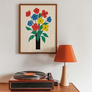 May include: A framed print of a colorful bouquet of red, blue, and yellow flowers in a black vase. The print is on a white background and is framed in a brown wood frame. A vintage record player and a lamp with an orange lampshade are in the foreground.