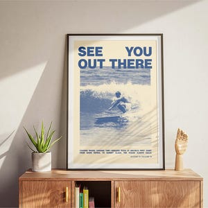 K&ouml;nnte beinhalten: Ein gerahmtes Poster mit einem blauen und wei&szlig;en Bild eines Surfers, der auf einer Welle reitet. Das Poster tr&auml;gt den Text "See You Out There" in blauen Buchstaben. Der Text unter dem Bild lautet: "Chasing waves, chasing the endless piece of balmy first light from dawn patrol to sunset glass. The ocean always calls. Bloody thirsty?"