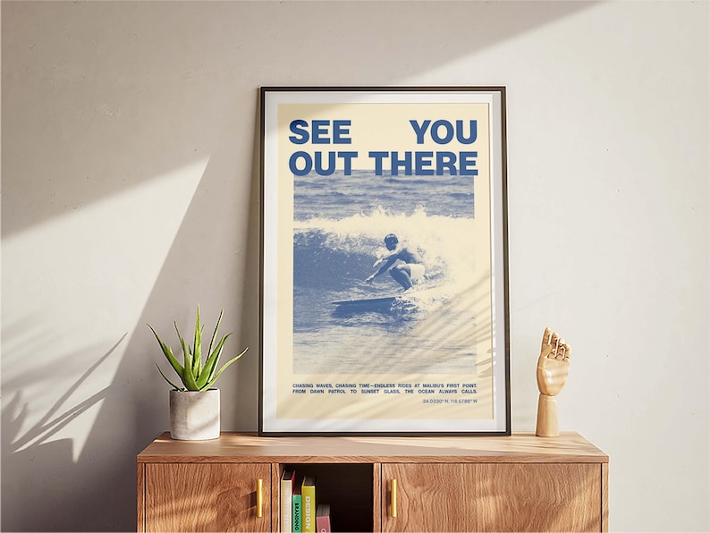 K&ouml;nnte beinhalten: Ein gerahmtes Poster mit einem blauen und wei&szlig;en Bild eines Surfers, der auf einer Welle reitet. Das Poster tr&auml;gt den Text "See You Out There" in blauen Buchstaben. Der Text unter dem Bild lautet: "Chasing waves, chasing the endless piece of balmy first light from dawn patrol to sunset glass. The ocean always calls. Bloody thirsty?"