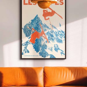May include: Poster with a vintage ski theme. The artwork features skiers in orange against a blue mountain backdrop. The text "LES'S" and "LA MONTAGNE CA VOUS GAGNE" are visible. The poster is framed and hangs above an orange leather sofa.