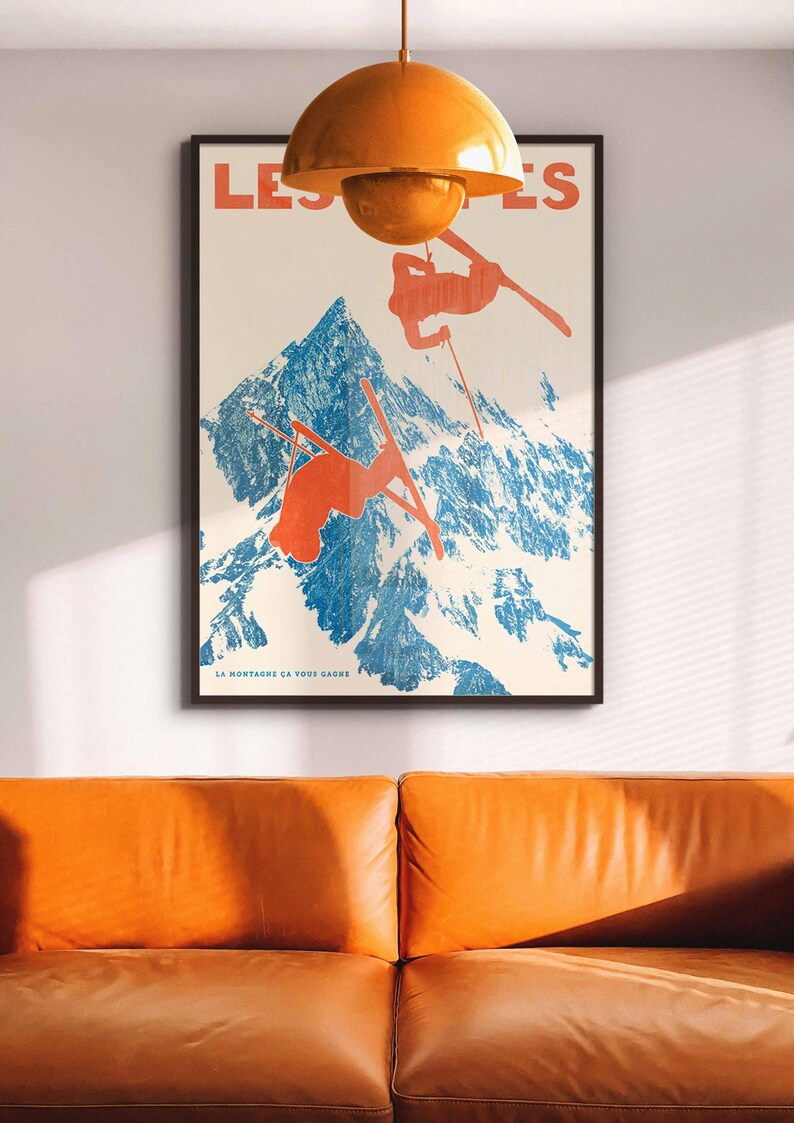 May include: Poster with a vintage ski theme. The artwork features skiers in orange against a blue mountain backdrop. The text "LES'S" and "LA MONTAGNE CA VOUS GAGNE" are visible. The poster is framed and hangs above an orange leather sofa.