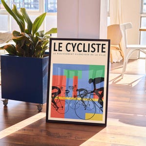 May include: Framed art print titled "LE CYCLISTE" featuring cyclists on bicycles with colorful geometric shapes in the background. The artwork is in a black frame and is displayed on a wooden floor.
