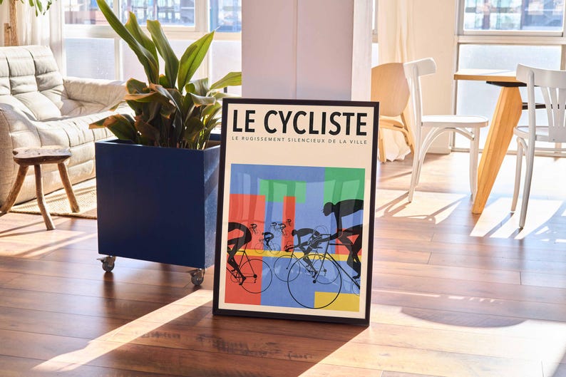 May include: Framed art print titled "LE CYCLISTE" featuring cyclists on bicycles with colorful geometric shapes in the background. The artwork is in a black frame and is displayed on a wooden floor.