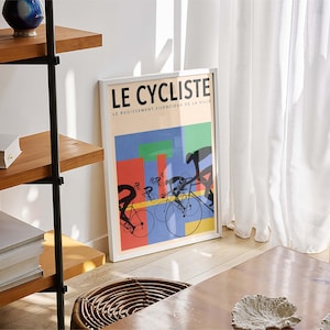 May include: A framed poster titled "LE CYCLISTE" with a colorful design featuring cyclists on bicycles. The poster is leaning against a white wall near a window with white curtains. A wooden shelf and a woven stool are also visible.