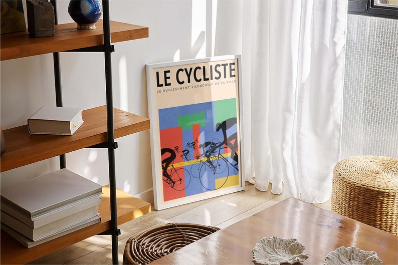 May include: A framed poster titled "LE CYCLISTE" with a colorful design featuring cyclists on bicycles. The poster is leaning against a white wall near a window with white curtains. A wooden shelf and a woven stool are also visible.