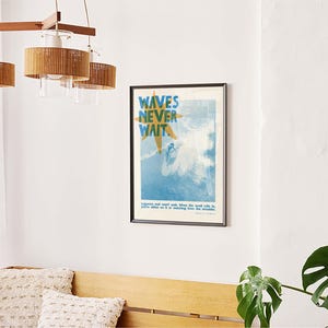 May include: A framed poster with the text "WAVES NEVER WAIT" in blue and yellow, depicting a surfer in the ocean. The poster is hanging on a white wall. A wooden bench with two decorative pillows is below the poster. A wooden and rattan chandelier hangs above.
