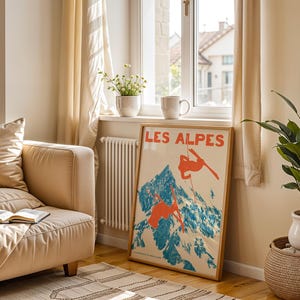 May include: A framed vintage poster titled "LES ALPES" depicts skiers on a mountain with blue and orange hues. The poster is propped against a wall near a window, a beige sofa, and a potted plant.
