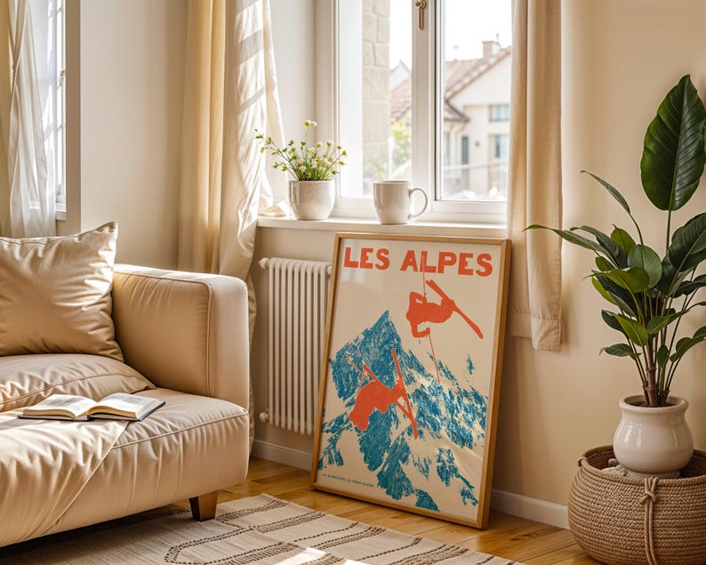 May include: A framed vintage poster titled "LES ALPES" depicts skiers on a mountain with blue and orange hues. The poster is propped against a wall near a window, a beige sofa, and a potted plant.