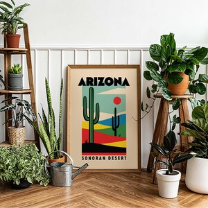 Może przedstawiać: Ramka z plakatem z tekstem "ARIZONA SONORAN DESERT" i kolorową ilustracją dw&oacute;ch kaktus&oacute;w i czerwonego słońca. Plakat jest otoczony doniczkowymi roślinami, w tym wężownicą, figowcem lirolistnym i gumowcem.