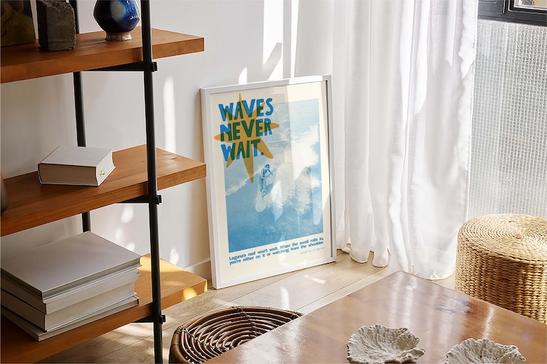 May include: A framed poster with the text "WAVES NEVER WAIT" in blue and yellow, depicting a surfer on a wave. The poster is leaning against a wall near a wooden table and a woven basket. A wooden shelf with books and decorative items is visible.