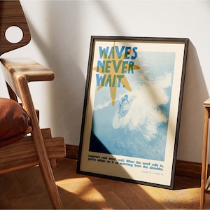May include: Framed poster with a surfing theme. The poster features the text "WAVES NEVER WAIT" in blue and yellow, with an illustration of a surfer riding a wave. The poster is in a black frame and leans against a wooden chair.
