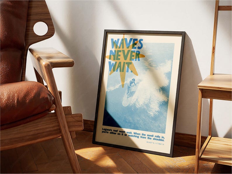May include: Framed poster with a surfing theme. The poster features the text "WAVES NEVER WAIT" in blue and yellow, with an illustration of a surfer riding a wave. The poster is in a black frame and leans against a wooden chair.