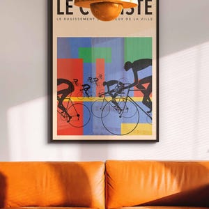 May include: Framed art print featuring a cycling race scene with silhouetted cyclists against a colorful abstract background. The artwork includes the text "LE CISTE" and "LE RUGISSEMENT S...EUX DE LA VILLE". The print is displayed above an orange leather sofa.