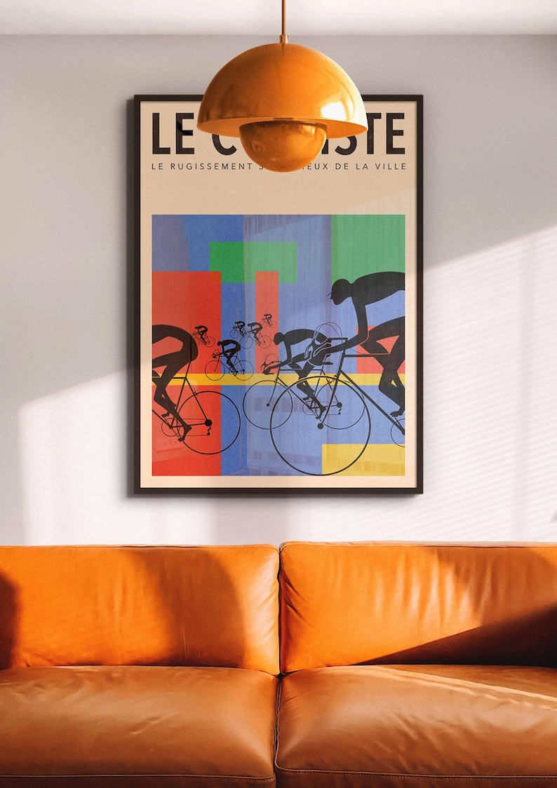 May include: Framed art print featuring a cycling race scene with silhouetted cyclists against a colorful abstract background. The artwork includes the text "LE CISTE" and "LE RUGISSEMENT S...EUX DE LA VILLE". The print is displayed above an orange leather sofa.
