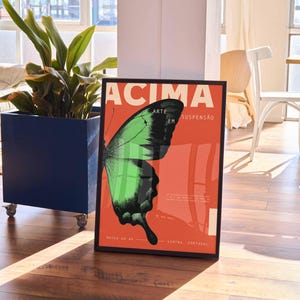 May include: A framed poster with a butterfly wing design in green and black on a red backdrop. The poster includes the word "ACIMA" and other text. The poster is displayed on a wooden floor.