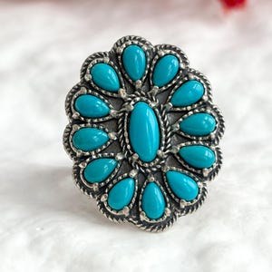 May include: A silver ring with a cluster of turquoise stones. The ring has an oval central stone surrounded by teardrop-shaped stones, all set in a detailed, textured setting. The design resembles a floral or sunburst pattern. The ring is a statement piece.
