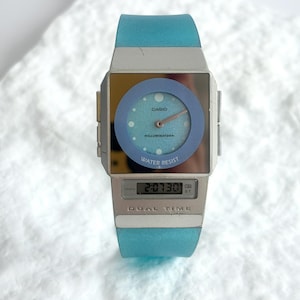 May include: A Casio Illuminator dual time watch with a light blue translucent silicone strap and a silver-toned rectangular case. The watch face features an analog dial with a blue circular bezel and a digital display window showing 2:07:30.