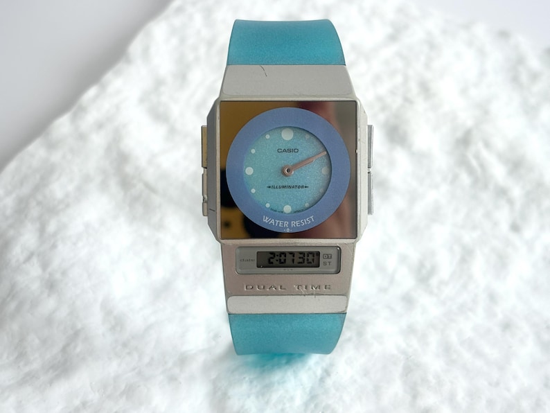 May include: A Casio Illuminator dual time watch with a light blue translucent silicone strap and a silver-toned rectangular case. The watch face features an analog dial with a blue circular bezel and a digital display window showing 2:07:30.