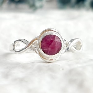 May include: A silver ring featuring a round, faceted, deep pinkish-red gemstone set in a smooth bezel. The band is crafted with an infinity-style design on both sides of the central stone, creating a continuous loop pattern that tapers toward the back.