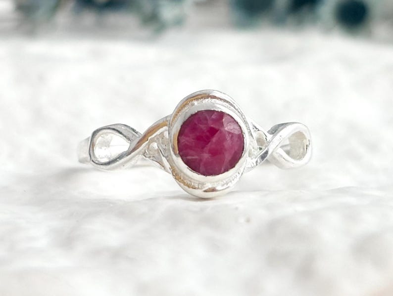 K&ouml;nnte beinhalten: Ein Silberring mit einem runden, facettierten, tief rosa-roten Edelstein in einer glatten Fassung. Die Ringschiene ist auf beiden Seiten des zentralen Steins mit einem Unendlichkeitsmotiv gestaltet, das ein durchgehendes Schleifenmuster bildet.