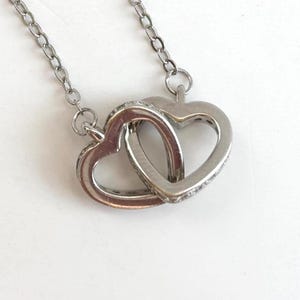May include: A silver-toned necklace featuring two interlocking heart-shaped pendants. The hearts are connected to a delicate link chain. The surface of the heart pendants is accented with small, clear, sparkling stones embedded along the outer edges.