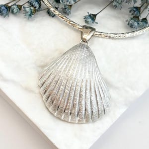 May include: A silver-toned seashell pendant suspended from a matching metallic omega chain necklace. The shell features a textured, ribbed surface with a metallic sheen, attached to the chain by a simple silver-toned bail at the top center.
