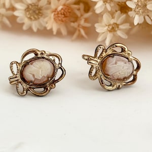 May include: A pair of vintage cameo earrings. Each earring features an oval cameo with a profile carving in a light peach colour, set in a gold-toned filigree frame. The earrings have a post-back closure and are displayed on a white surface.