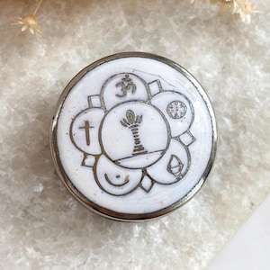 Puede incluir: Un colgante redondo de metal blanco con borde plateado. Presenta un conjunto de símbolos religiosos en contorno plateado, incluyendo una cruz, el símbolo Om y un diseño en forma de árbol. El fondo es una superficie clara y texturizada.