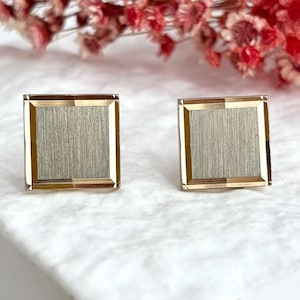 May include: A pair of square cufflinks with a gold-tone frame and a brushed silver-tone center. The cufflinks are displayed on a white surface with dried red flowers in the background. These accessories are suitable for formal wear.