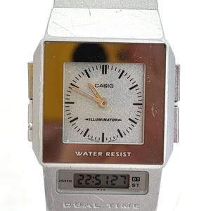 May include: Silver Casio digital watch with a square face and a digital display showing the time. The watch face has a silver background with black markings and gold hands. The watch is labeled "CASIO" and "WATER RESIST".