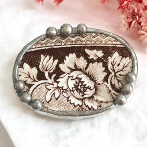 May include: An oval brooch featuring a piece of brown and white floral patterned ceramic set in a soldered silver-toned metal frame. The frame is decorated with small metal spheres along the top and bottom edges, creating a textured border.