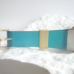 May include: A wristwatch featuring a rectangular silver-tone case and a light blue dial with a dark blue circular accent. The strap is a gradient of blue with a cream-colored buckle and a matching cream-colored sliding strap keeper.
