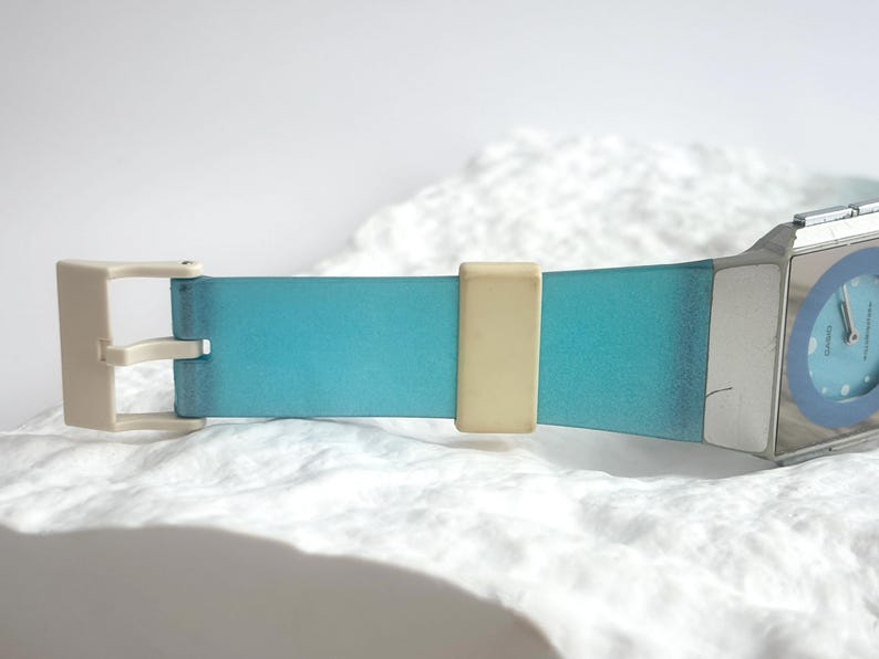 May include: A wristwatch featuring a rectangular silver-tone case and a light blue dial with a dark blue circular accent. The strap is a gradient of blue with a cream-colored buckle and a matching cream-colored sliding strap keeper.