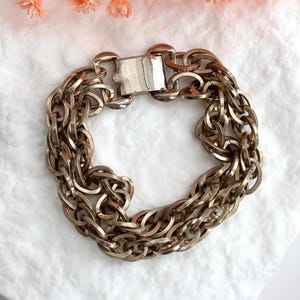 May include: A silver-toned chain bracelet with multiple interwoven strands. The bracelet features a rectangular clasp and a textured, metallic finish. The jewelry is displayed on a white surface with a soft, floral accent.