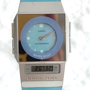 May include: A Casio dual time watch featuring a square silver-tone case with a light blue circular dial, analog hands, and a digital display window at the bottom. The watch face includes the text Casio, Illuminator, Water Resist, and Dual Time.